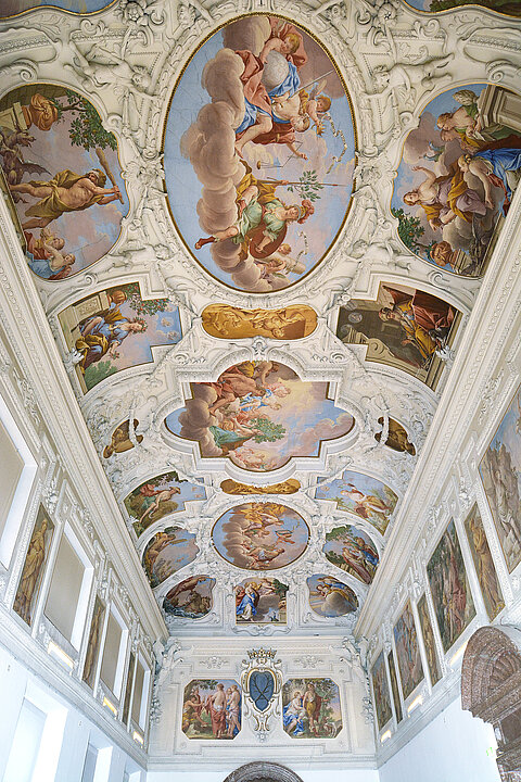 
The ceiling of a large hall decorated with white stucco and colorful paintings. The paintings show figures from legends and religions.