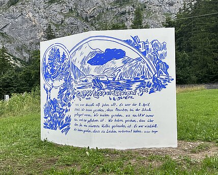 Auf einer Wiese steht eine weiße Skulptur wie eine Seite von einem Buch, in der Mitte ist diese rechteckige Wand leicht geknickt. Darauf ist in Blau eine Zeichung mit den Worten: Gruss vom Leopoldsteiner See bei Eisenerz. Ich war damals elf Jahre alt. Es war der 8. April 1945. Wir haben gesehen, dass Menschen bei der Schule gelagert waren. Wir haben gesehen, wie ein LKW immer hin und her gefahren ist. wir haben gesehen, dass über dem See eine schwarze Wolke gestanden ist. Es war windstill. Wir haben gesehen, dass sie Leichen verbrannt haben. Oskar Berger