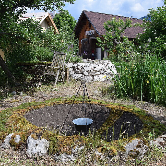 Am Foto ist ein Garten mit Gartenstuhl, Steinmauer, Pflanzen, Bäumen. Im Hintergrund sieht man zwei Gebäude. Im Vordergrund ist ein Loch in der Erde, in dem in der Mitte ein Kessel auf einem Dreibein hängt. Rund um das Loch liegt Moos.