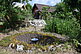 Am Foto ist ein Garten mit Gartenstuhl, Steinmauer, Pflanzen, Bäumen. Im Hintergrund sieht man zwei Gebäude. Im Vordergrund ist ein Loch in der Erde, in dem in der Mitte ein Kessel auf einem Dreibein hängt. Rund um das Loch liegt Moos.