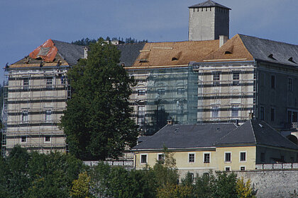  Photograph of Trautenfels Castle. There is scaffolding on the castle walls. On the roof there are workmen who are redoing the roof. Parts of the roof are already light brown with new shingles, parts are still dark brown with old shingles.