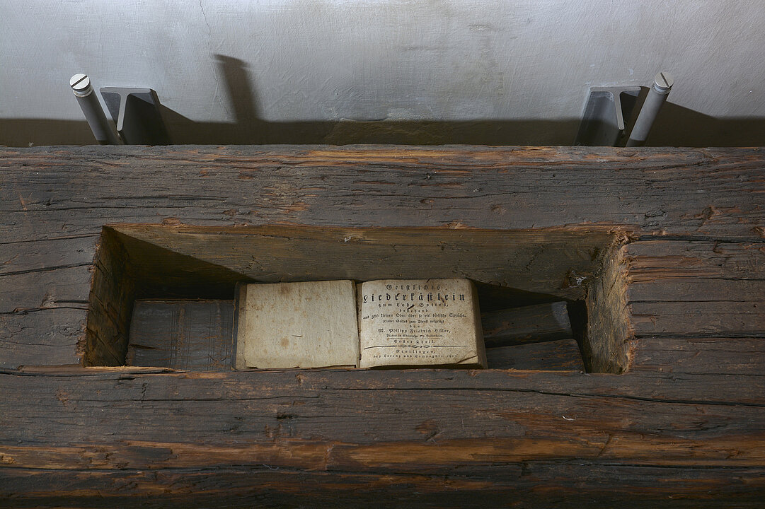 Brauner Holzbalken. In der Mitte ist ein Quader aus dem Balken ausgeschnitten, darin liegt ein kleines aufgeschlagenes Buch.
