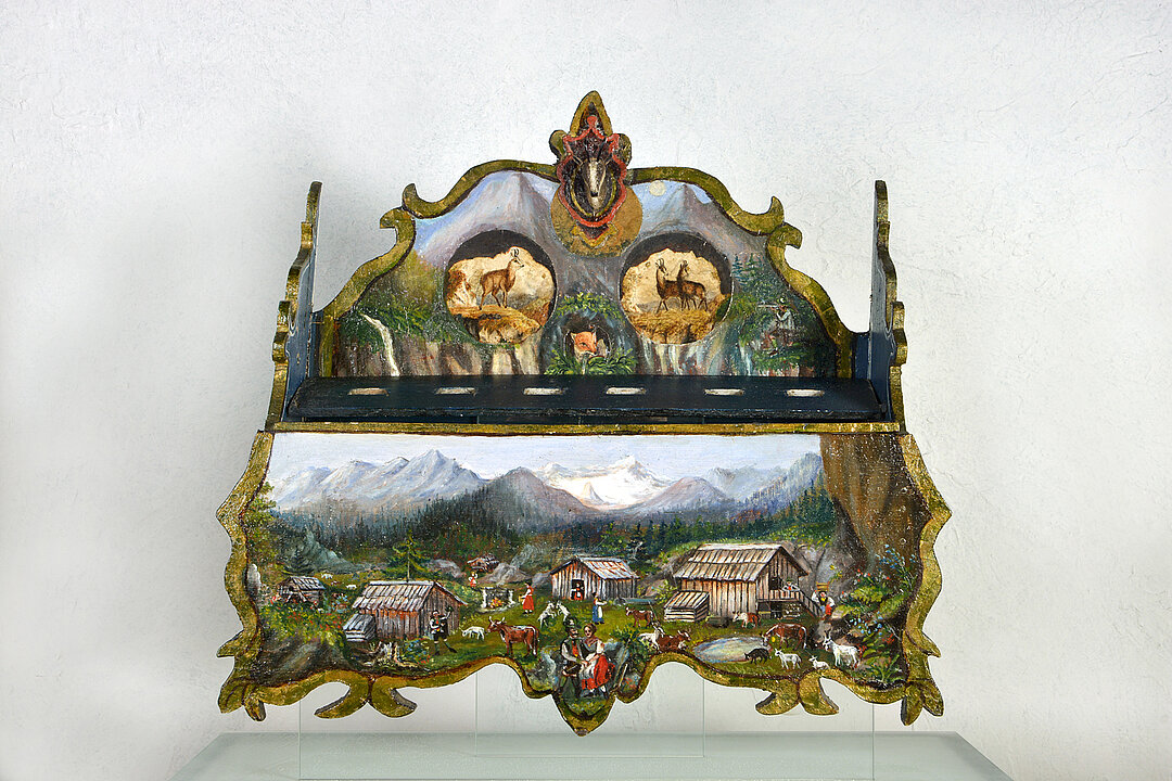 Hanging device for spoons. There are six holes on a shelf into which spoons can be inserted. Shelf is colorfully designed. You can see alpine huts in front of mountains and chamois and ibex.