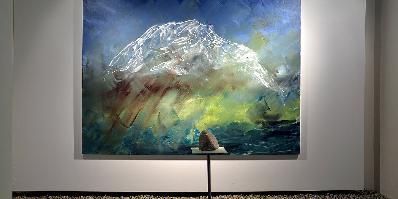 Ein Kalstein-Felsbrocken liegt vor einem Gemälde auf einem Podest. Auf dem Gemälde ist der Berg Grimming zu sehen.