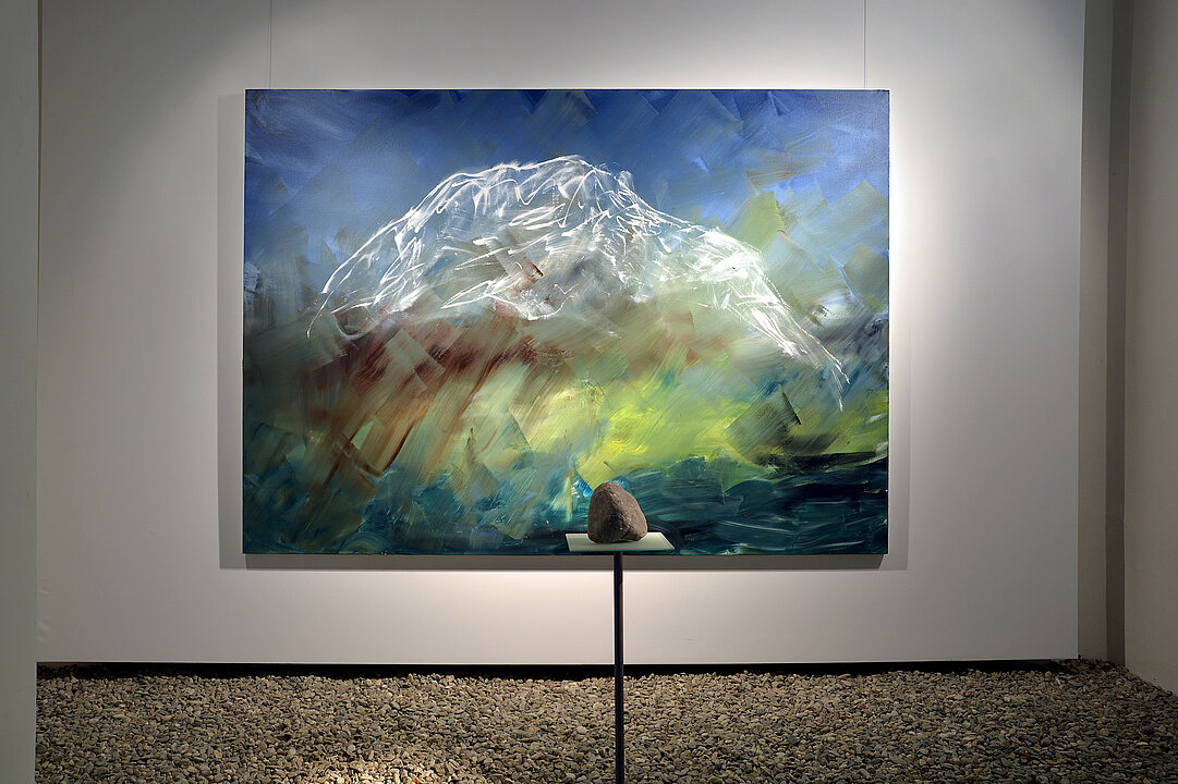 A limestone rock lies on a pedestal in front of a painting. The painting shows the mountain Grimming.