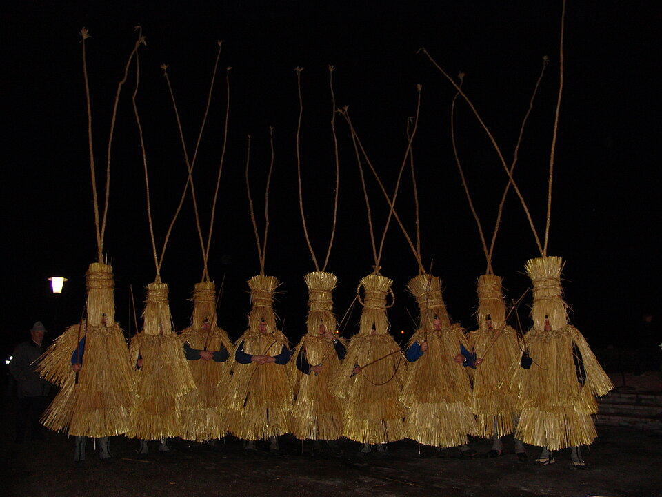 Foto von neun Menschen, die die Verkleidung "Schab" tragen. Sie tragen ein ganz in Stroh gekleidetes Gewand. Dazu kommen vier bis fünf Meter hohe Hörner.
Es ist NAcht.