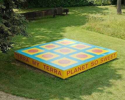 Die Skulptur ist eine rechteckige Plattform mit der Aufschrift "TERRA MY TERRA/PLANET SO SWEET/I CAN FEEL YOU/UNDER MY FEET". Die Plattform ist bemalt mit geometrischen Formen in blau, gelb und rot.