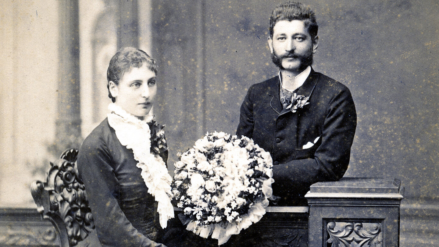 An old wedding photo. The bride wears a dark dress and holds a large bouquet of flowers. The man is standing behind there and is wearing a dark suit. They both smile very slightly.