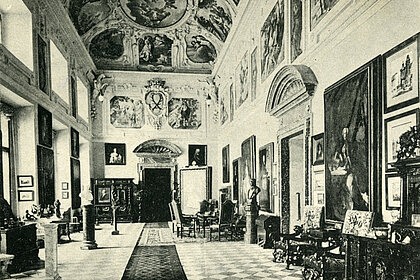  Photograph of a large hall. Many paintings hang on the walls. There are statues and large wooden furniture in the room.