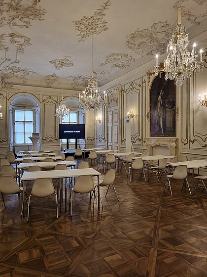 In Prunkraum 207, tables with chairs are arranged facing the stage. There is a speaker's podium and a large screen on the stage.