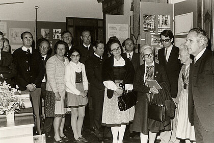  Black and white photograph of a group of people. They are standing and looking at a man who is speaking. Many are wearing traditional costumes, there is one child.