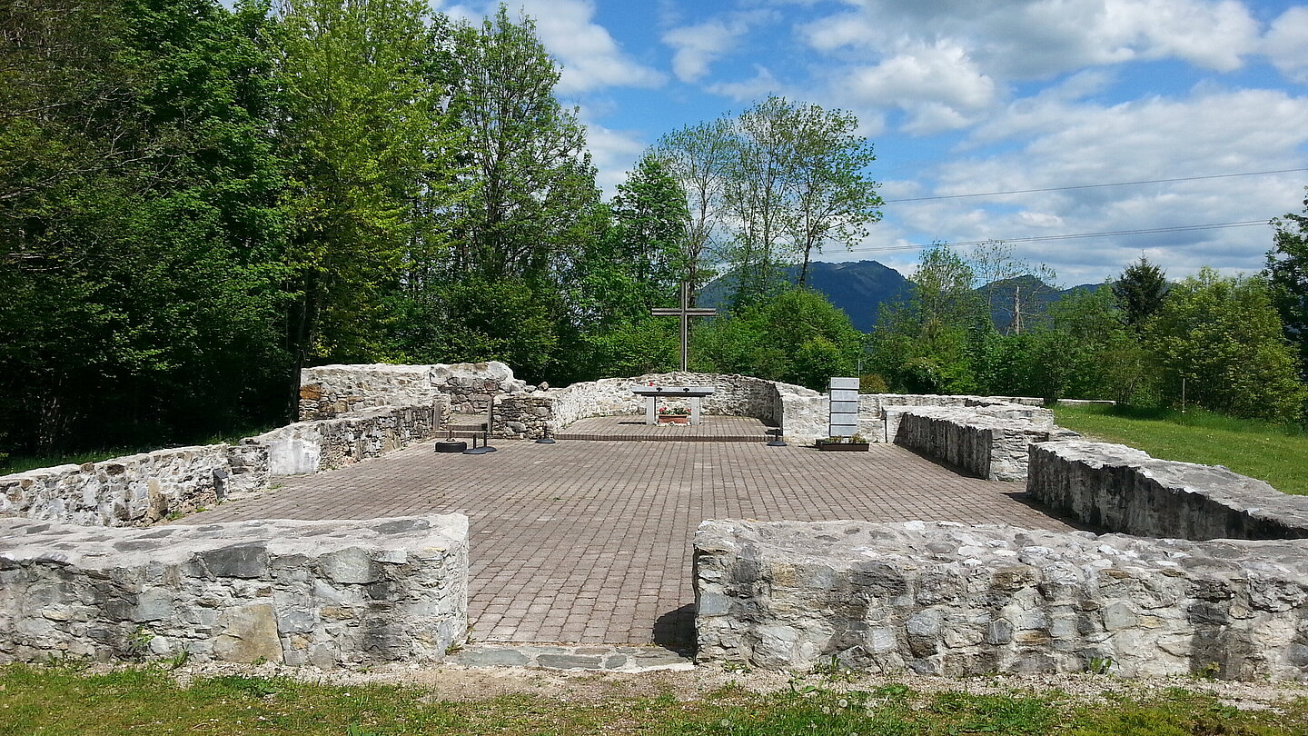 
Photograph of a church ruin. Only the foundation walls are still standing. In the background are trees and a blue sky with clouds.