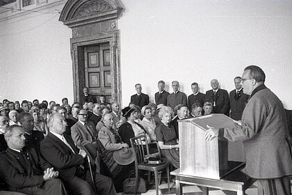 A black and white photograph. On the right of the picture is a man giving a speech. On the left, adults are sitting and standing and listening to the speaker. Some of the audience are wearing traditional clothes.