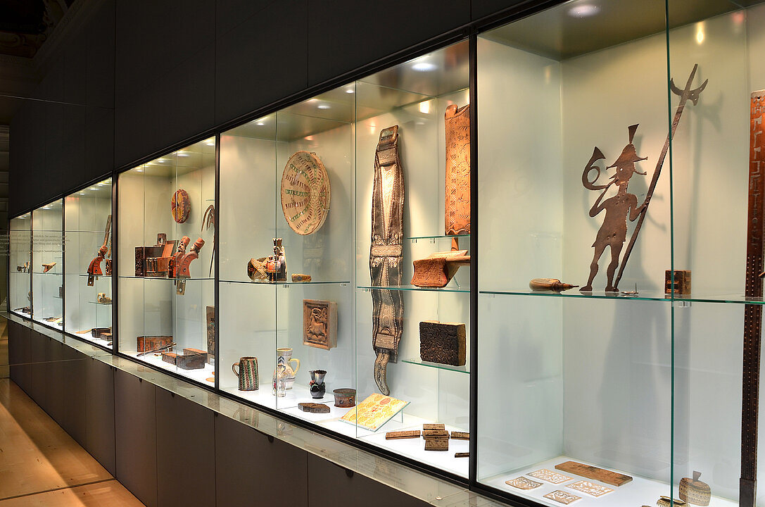 Exhibition view „Objects of Beauty" Photo of a long glass display case. In it lie various objects. These all have a use e.g. a bowl or a jug, but are all also specially decorated.