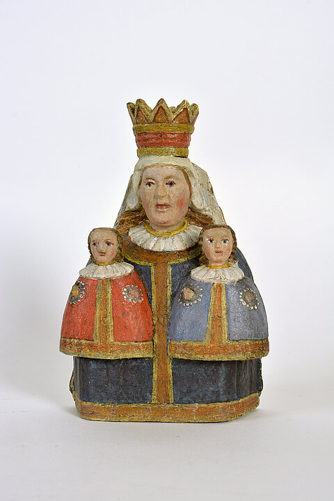 Holzfigur einer Frau mit Krone, die auf jeder Seite ein kleines Kind hat. Die Figur ist bemalt. Ein Kind hat einen blauen, ein anderes einen roten Mantel.