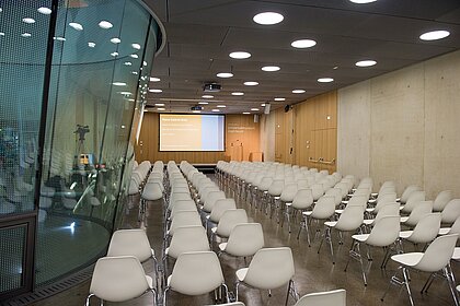 In the auditorium, chairs are arranged in rows facing the stage. A speaker's podium and a screen are projected onto the stage. A camera stands at the edge and films towards the stage.