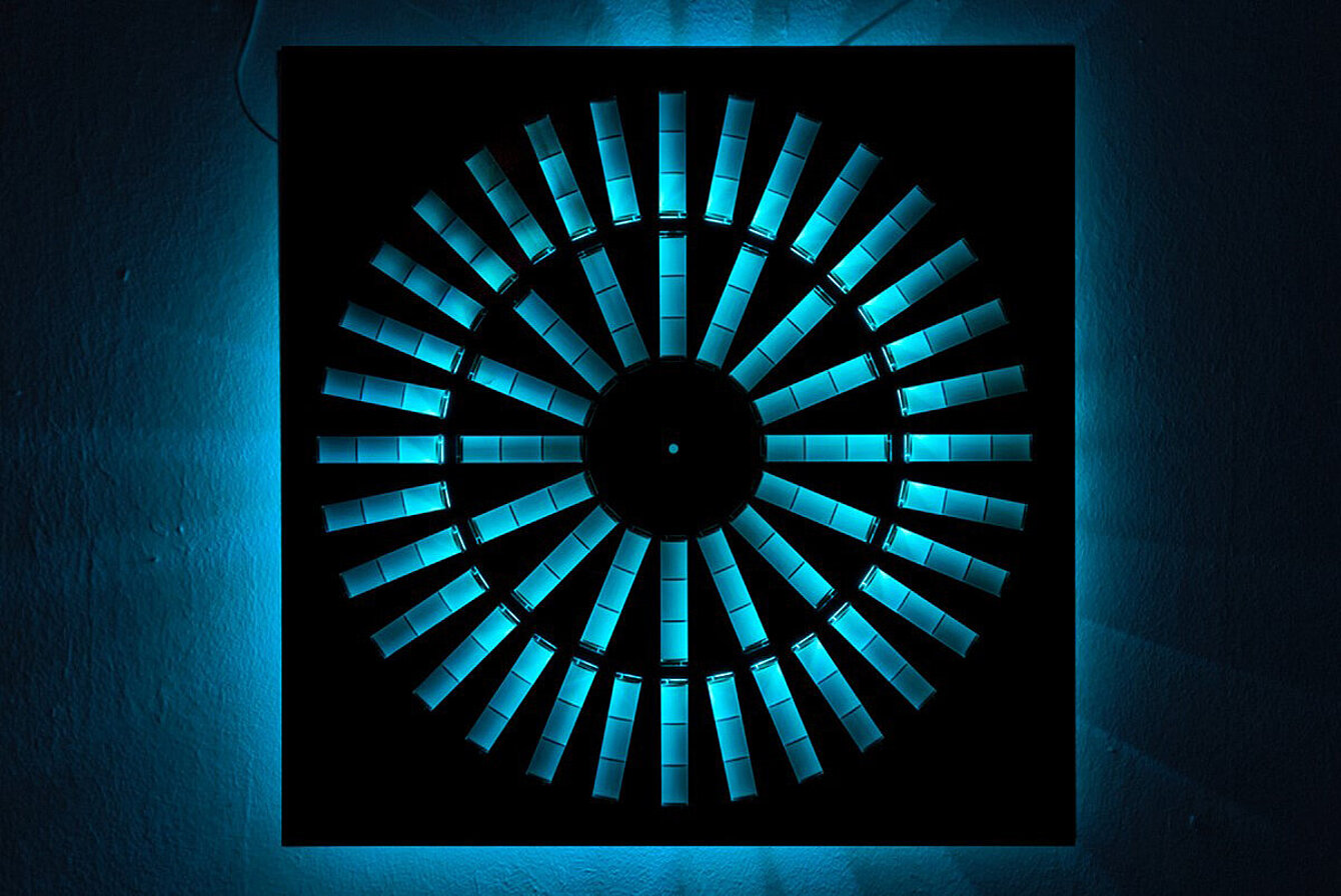 Photo of an art installation: circular rectangles are cut out of a black plate. Behind it is a blue light that falls through the cutouts. 