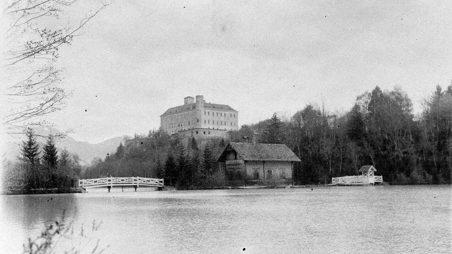 Black and white photograph of Trautenfels Castle. In the foreground is a lake. The sky is cloudy.
