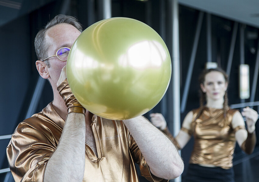 Footage of a performance in the Needle at the Kunsthaus Graz. In an extreme close-up and cropped shot, a man blows up a golden balloon. Another performer can be seen out of focus in the background.