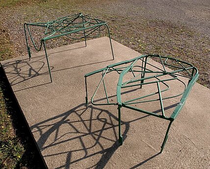 The two-part sculpture made of bent and green-painted metal struts resembles two stools in shape and rests on a concrete base.