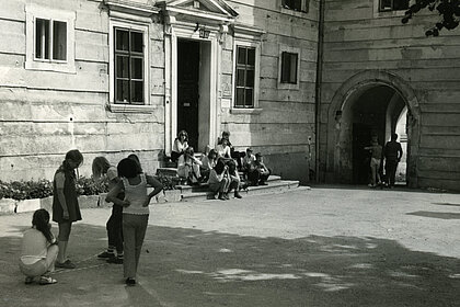 A black and white photograph of young people, in a courtyard. One group is in the foreground on the left, another continues to sit on a staircase.