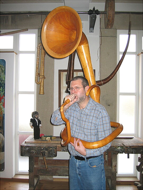 Ein Mann bläst in ein Horn aus Holz. Das Horn windet sich um seinen Oberkörper. Der Schallbecher ist einen Meter über seinem Kopf.