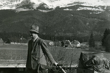  A man with a hat pulls a handcart on which wrapped objects lie. In the background one sees mountains and forests.