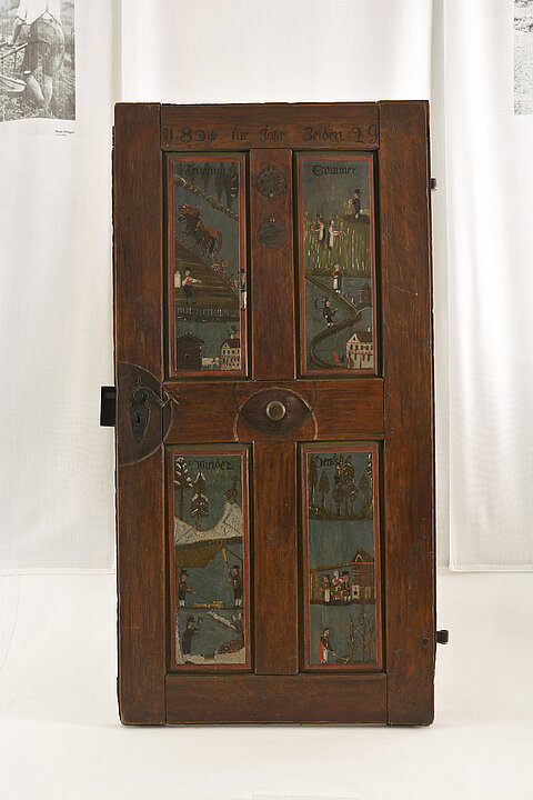 Eine Holztüre wird auf einem weißen Sockel ausgestellt. Die Türe ist mit dunklen Farben verziert, die Szenen aus den vier Jahreszeiten zeigen.