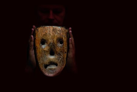 Das Foto ist fast komplett schwarz. Nur zwei Hände sind zu erkennen, eine Maske aus Holz halten. Von der Maske ist nur die Innenseite zusehen. Mit Löchern für einen Mund, zwei Augen und die Nase.