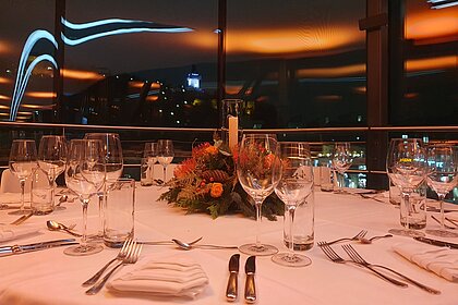 In the foreground is a covered round table. In the background, orange lights are reflected in the glass façade of the Needle. The clock tower on the Schlossberg can be seen.