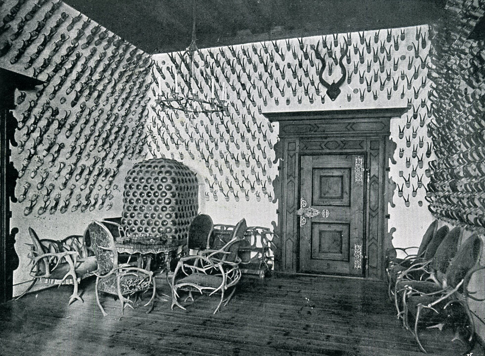 Old photograph of a room. Antlers hang from top to bottom on the walls. In the left corner there is a stove in front of which you can see pieces of furniture made from antlers.