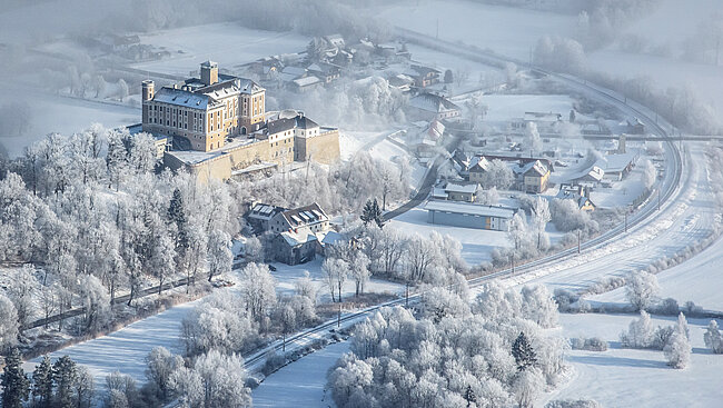 Trautenfels Castle photographed from the air. There is snow everywhere. Railroad tracks run along in front of the castle. Along the railroad line there are houses and trees.