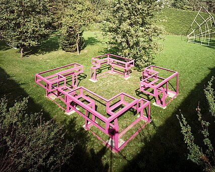 The sculpture is a large walk-in structure made of pink painted metal. The ground plan is square, with an "entrance" on each side.