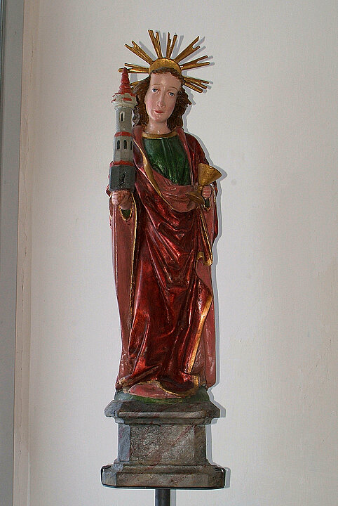 Holzstatue von einer Frau mit rotem Gewand und goldenem Heiligenschein. In der Hand hält sie einen kleinen grauen Turm mit rotem Spitzdach.
