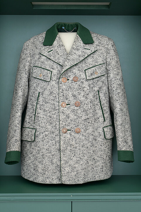 Foto von einem Mantel, der in einer Vitrine hängt. Der Mantel ist aus grauem Loden und hat einen grünen Kragen. An der Vorderseite befinden sich drei Reihen doppelte Knöpfe.