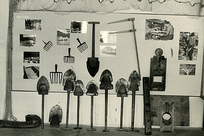 Black and white photograph of a wall. Various museum objects (mainly shovels) and photographs are attached to it.