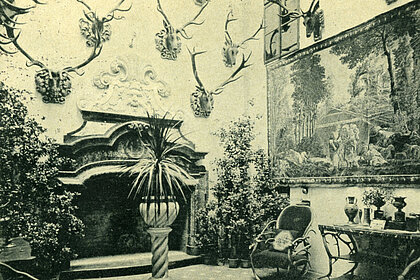 photograph of a room with a fireplace. There are antlers and a painting on the wall. There are plants everywhere