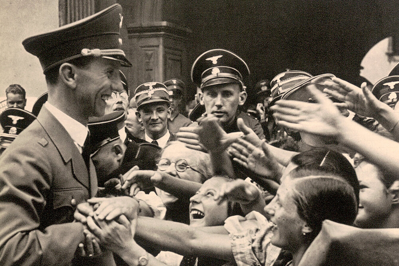 Schwarz-weiß Fotografie von eine Mann (Joseph Goebbels) in Uniform mit militärischer Mütze. Rechts von ihm stehen eine jubelnde Kindern. Sie strecken ihm die winkenden Hände entgegen oder halten seine Hand.