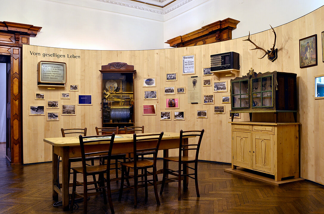 Photo of an exhibition room. The walls are covered with wood, on them hang many small pictures and photographs. In the middle of the room is a table with six chairs.